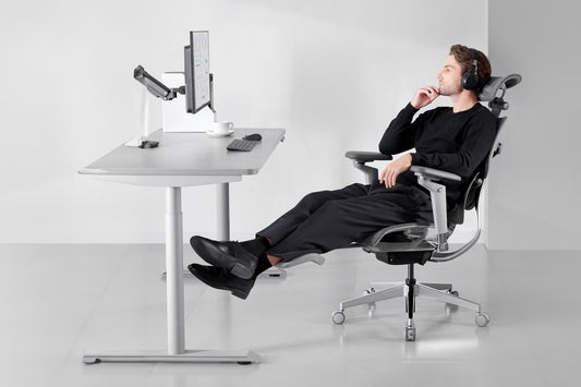 Man relaxing in a Hinomi ergonomic chair with footrest extended while working at a standing desk in a modern office.